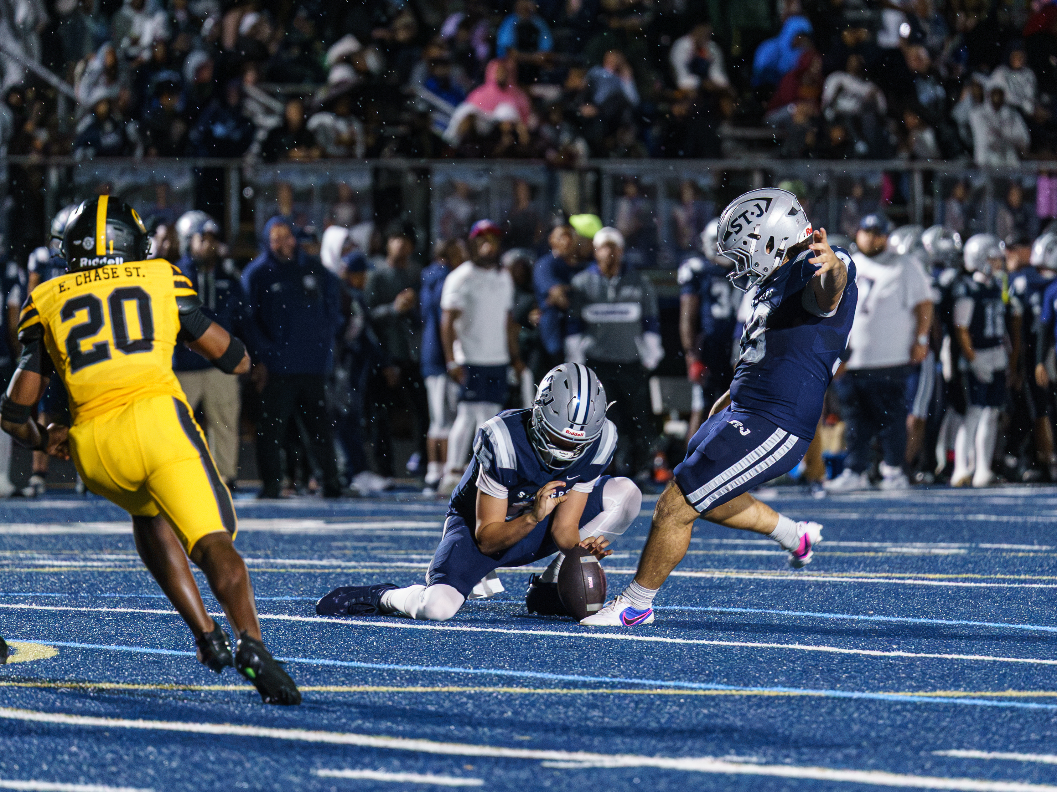 A St. James player kicks the football as a holder and an opposing player appear nearby.